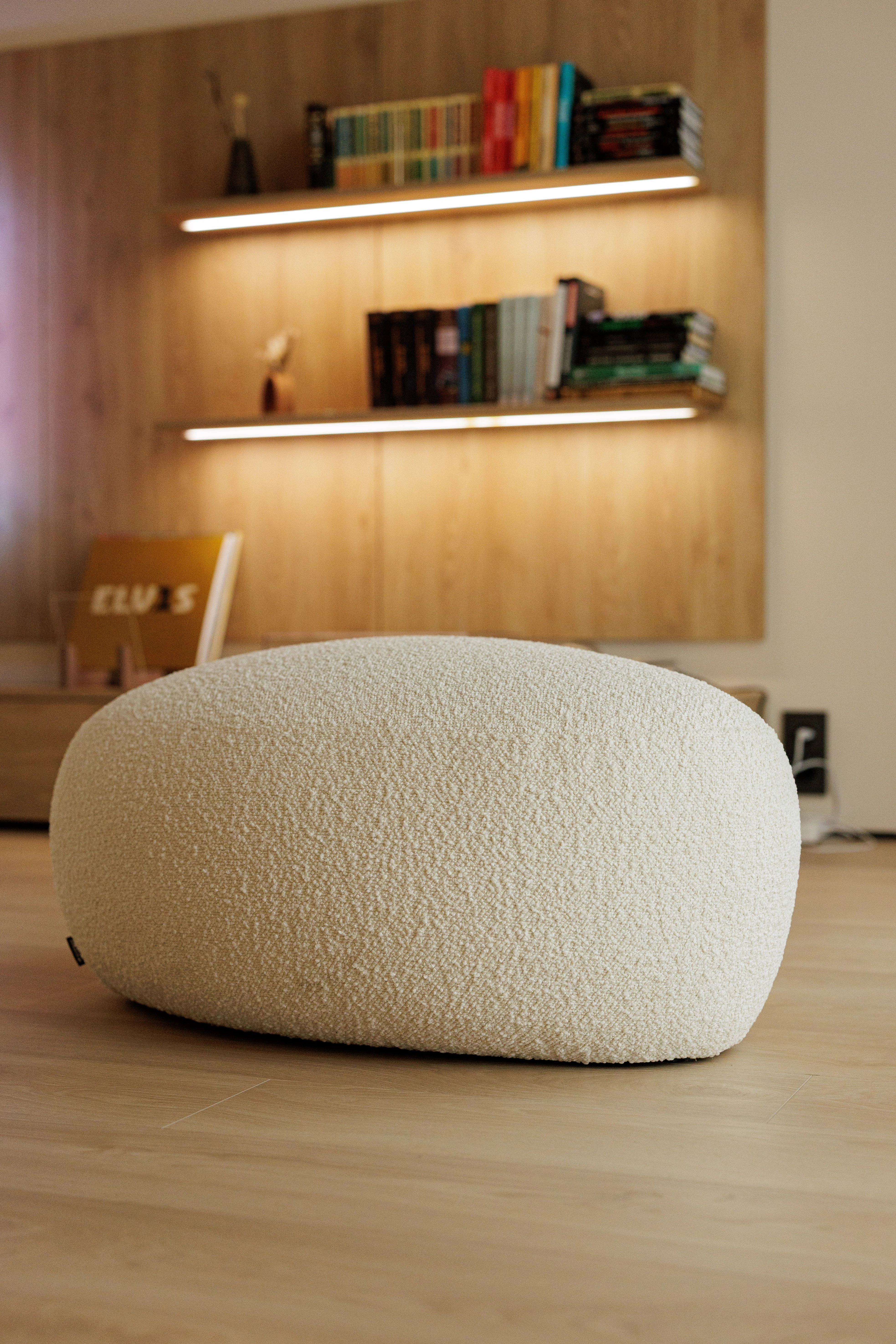 Cream bouclé Chios pouf in a warm apartment with a lit wood bookshelf behind