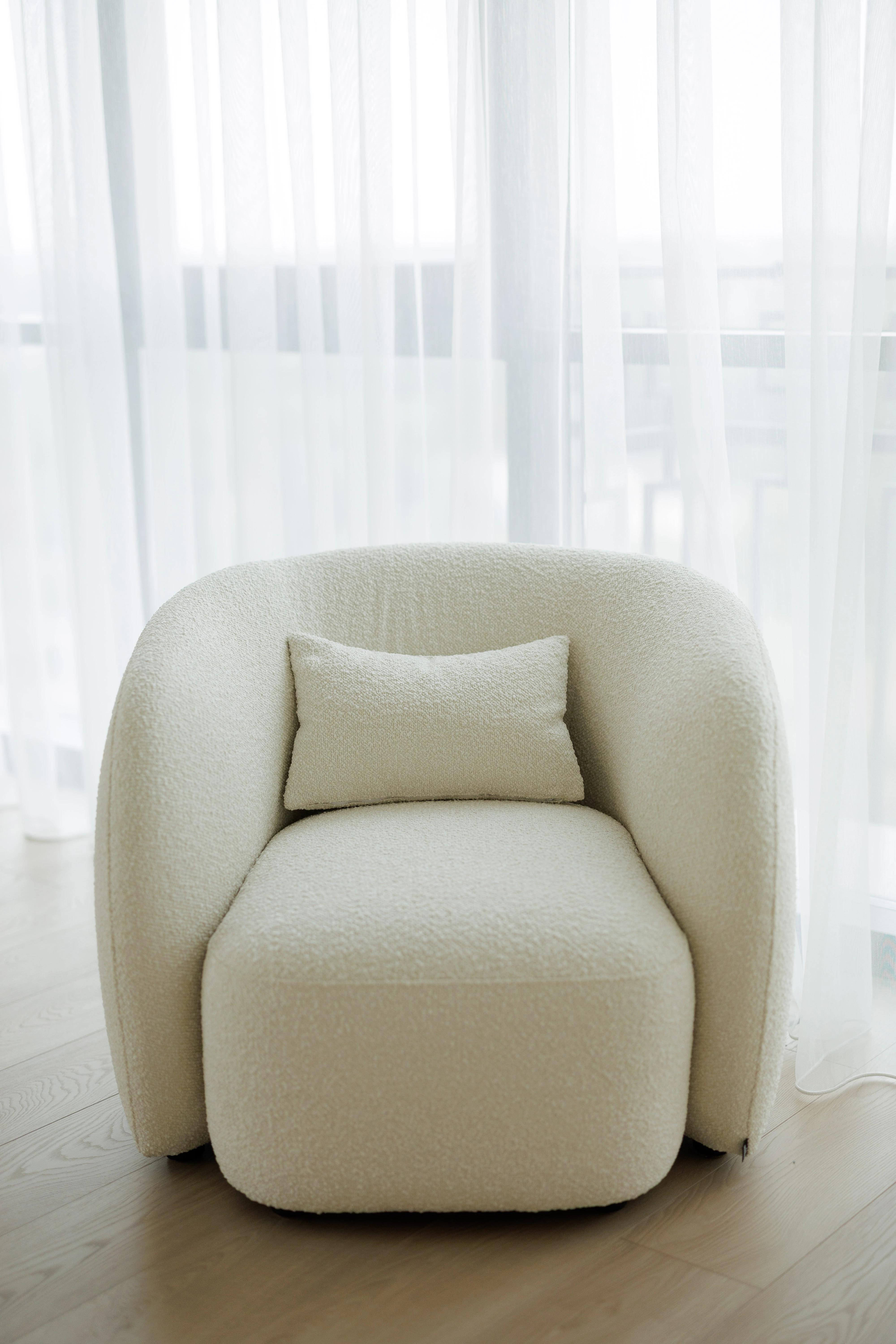 Cream boucle Nuba armchair alone, backlit by a wall of sheer white curtains on an oak floor