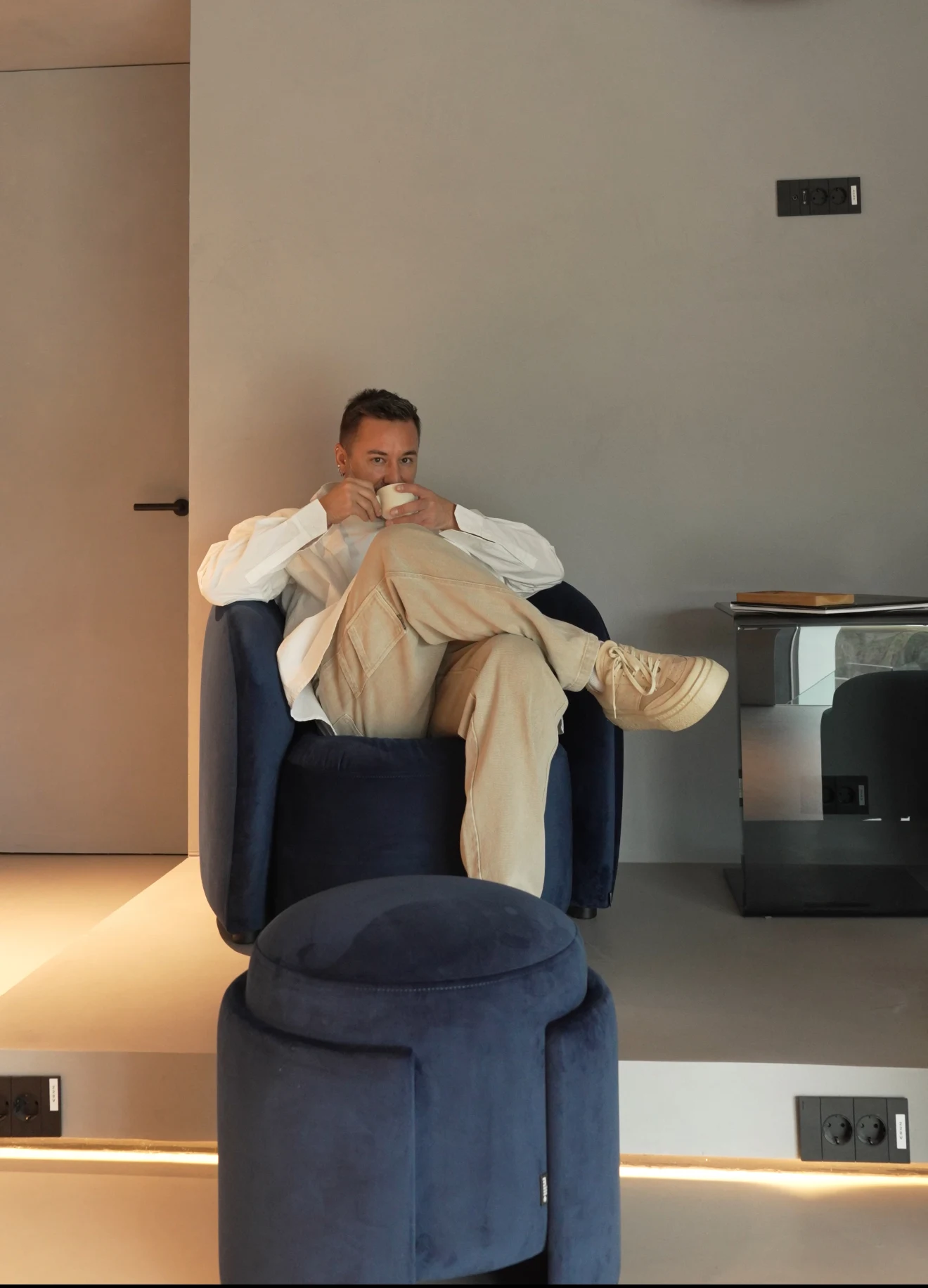 Eden pouf in navy velvet beside a navy armchair where a man sits with a coffee