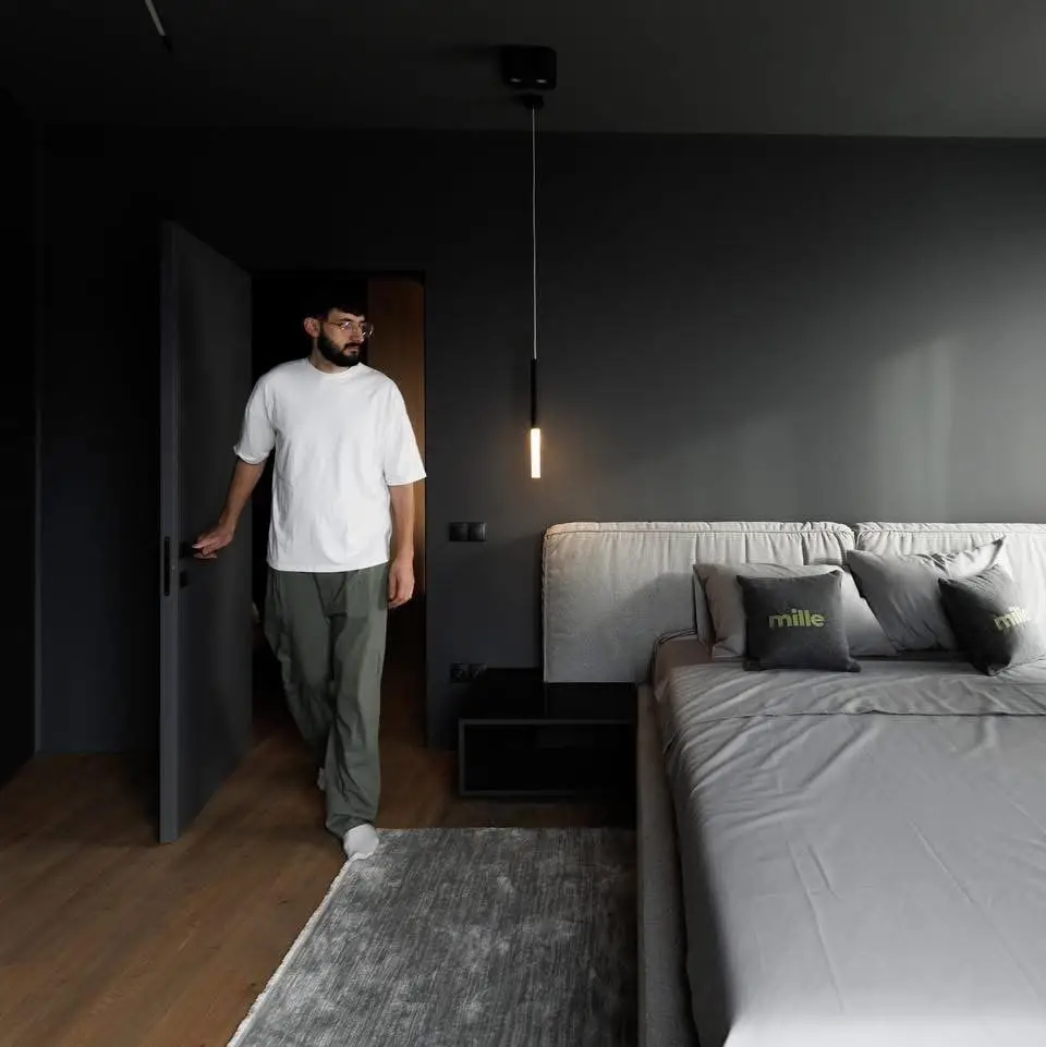 Mid-grey Palermo in a dark bedroom with a man in a white tee walking through the doorway, Mille pillows on the bed