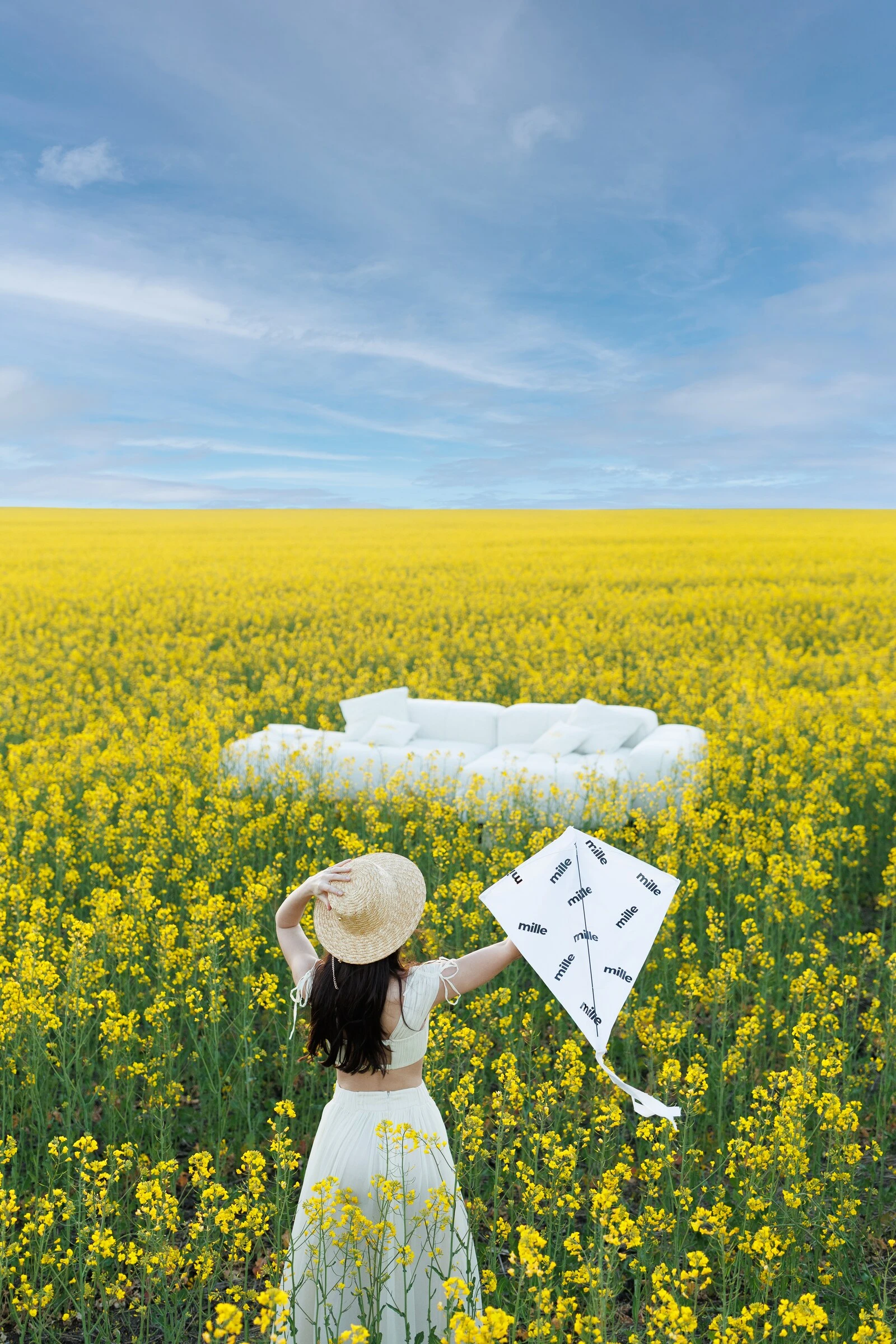Woman in white with a straw hat holding a Mille kite, cream Nami in the canola field behind