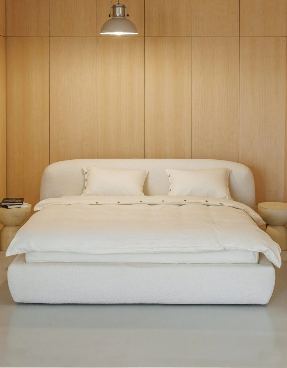 Cream boucle Belagio bed centered in a warm wood-paneled bedroom under a single industrial pendant lamp, two stone side tables flanking the bed