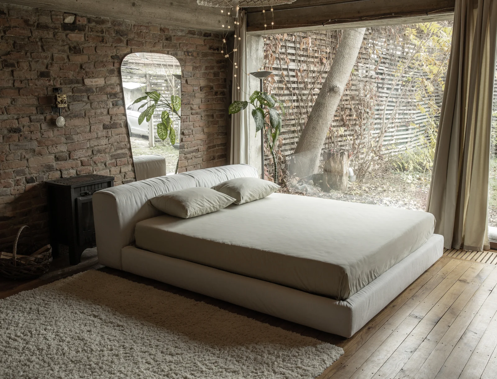 Cream Soho 900 Marmaris bed in a rustic brick loft with fairy lights overhead, a vintage mirror, a plant by a large window opening onto a sunny garden