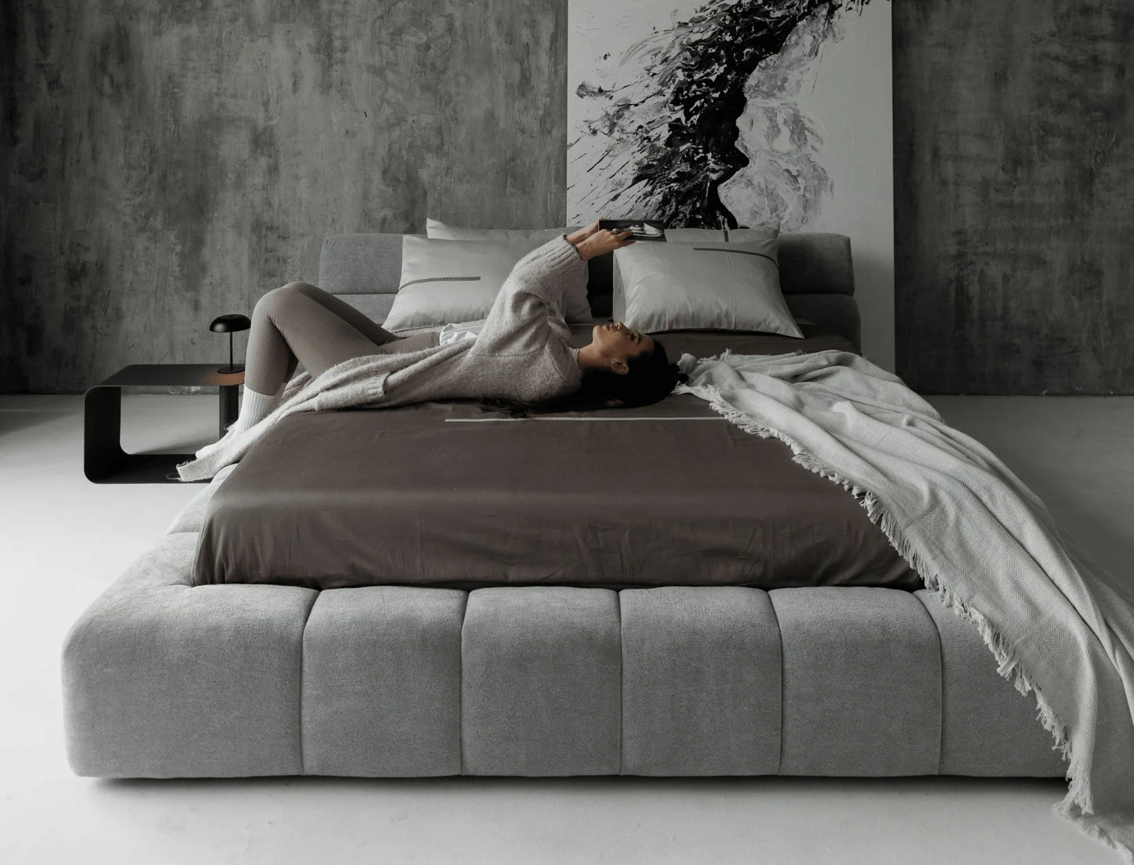 Mid-grey velvet Nao bed in a concrete-wall bedroom with a sumi-e ink art panel, model in cream loungewear reclining diagonally across it
