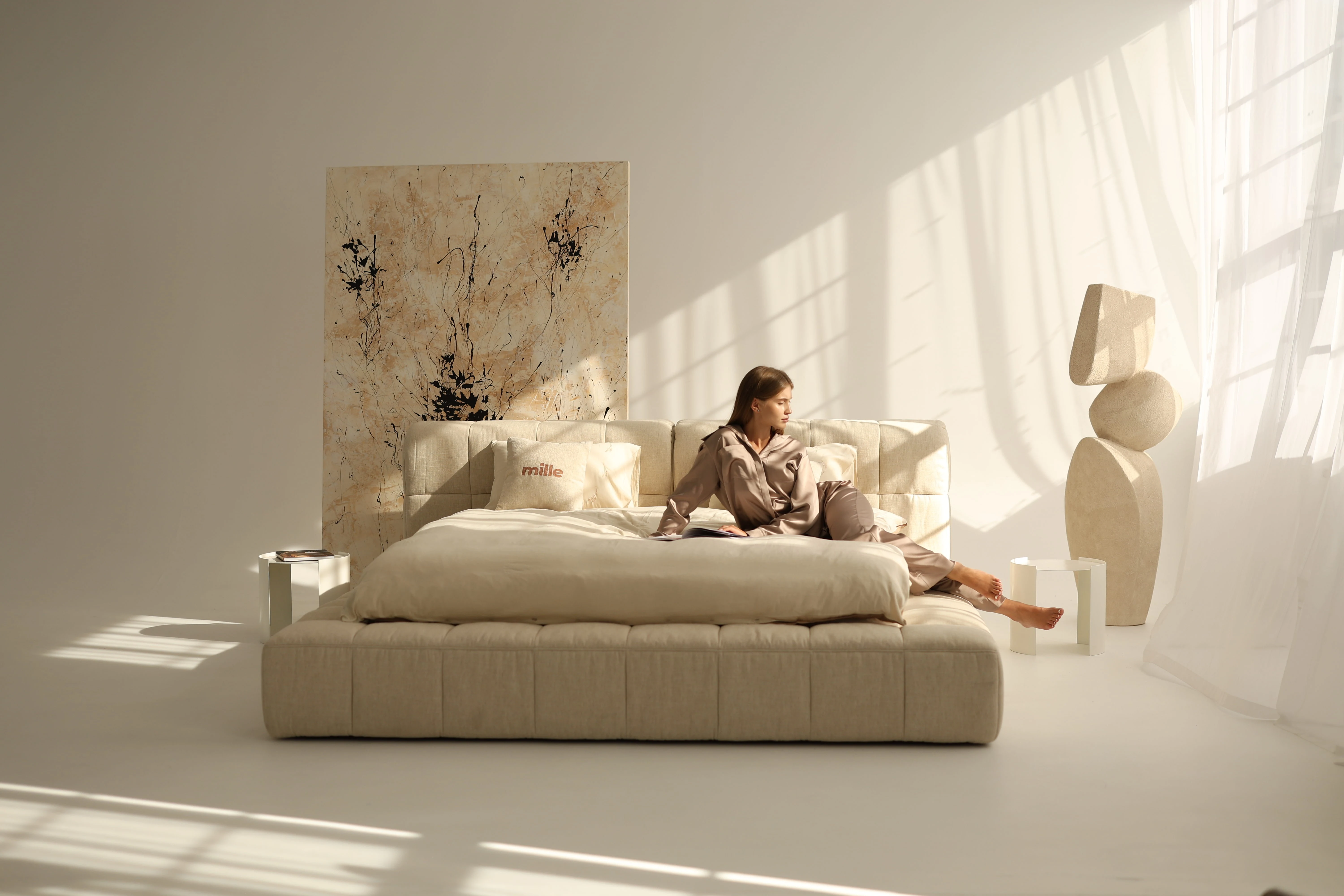Sandy linen-channelled Nao bed in a warm sunlit loft with an abstract art panel and sculptural pieces nearby, model in silk loungewear seated on the bed