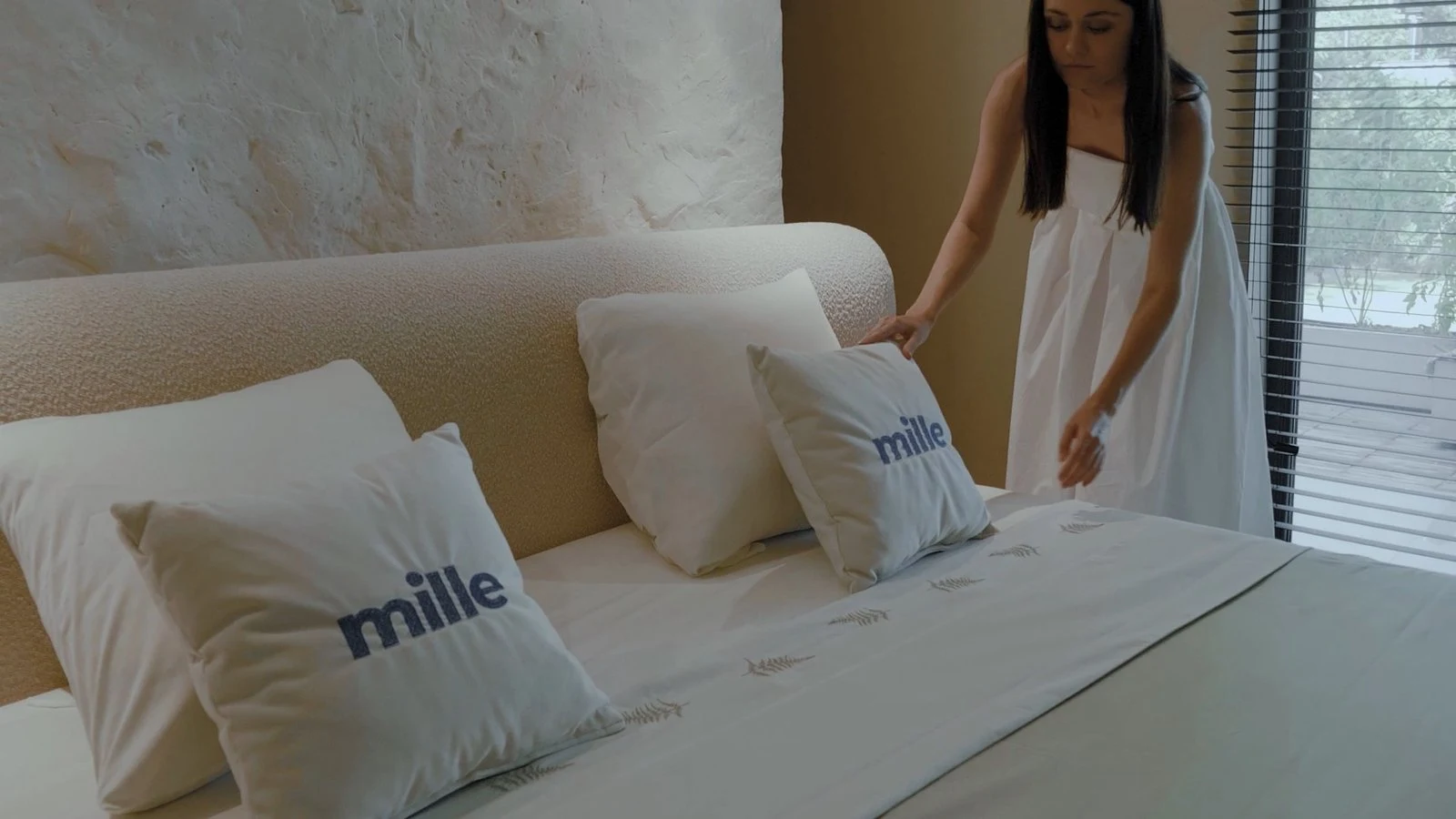 Cream Copenhagen 900 boucle Fresco bed in a textured-plaster-wall bedroom with a woman in a white dress arranging Mille-branded pillows on the bed