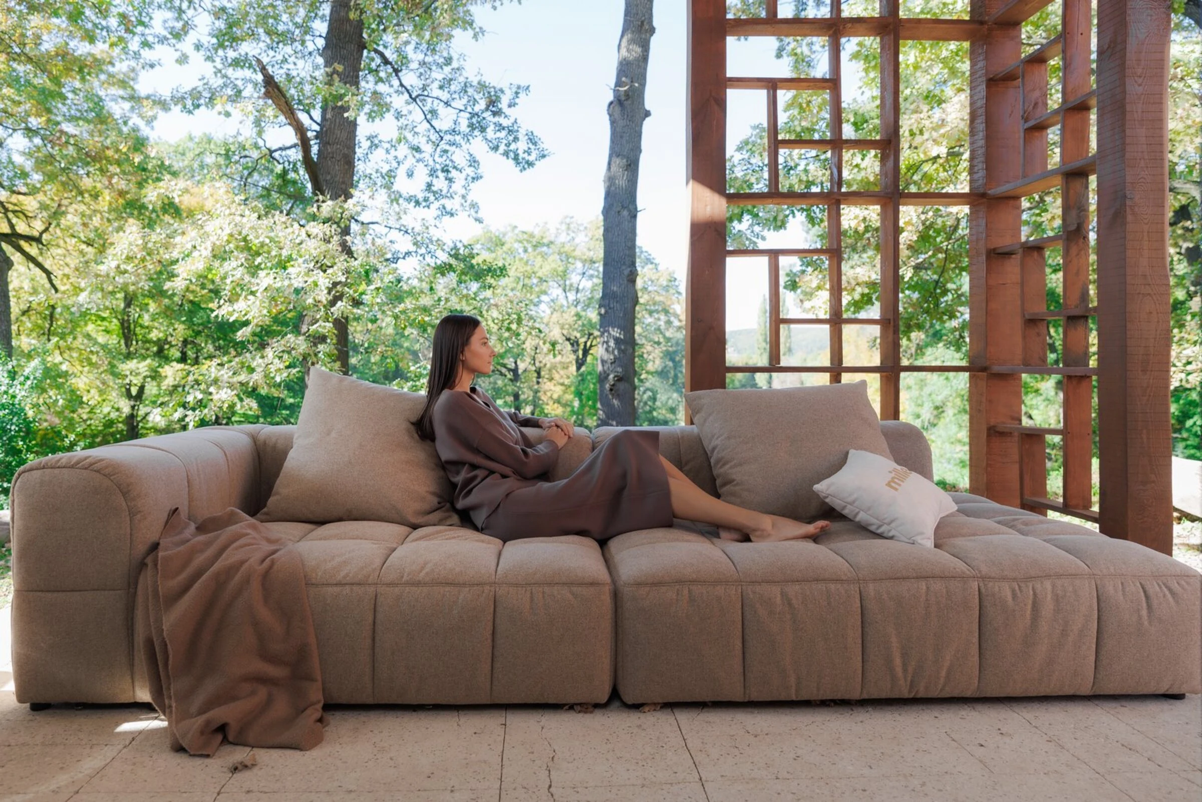 Tan oat Soft on a stone-and-wood terrace with mountain trees, model reading on the cushions